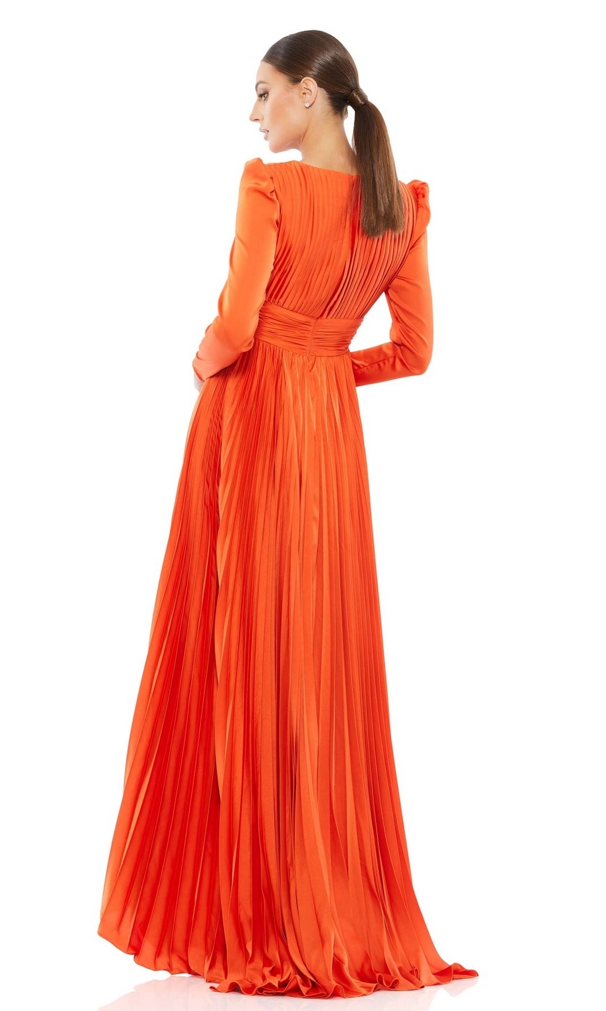 A woman with a low ponytail models the Mac Duggal Long Formal Dress 26542, an orange pleated gown with long sleeves and a floor-length A-line silhouette. She stands facing away, showcasing the gown’s elegant back detail.